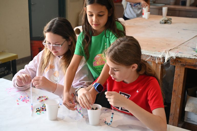 teenagers making friendship bracelets and girl in green shirt putting a bracelet on a girl in a red shirt's wrist
