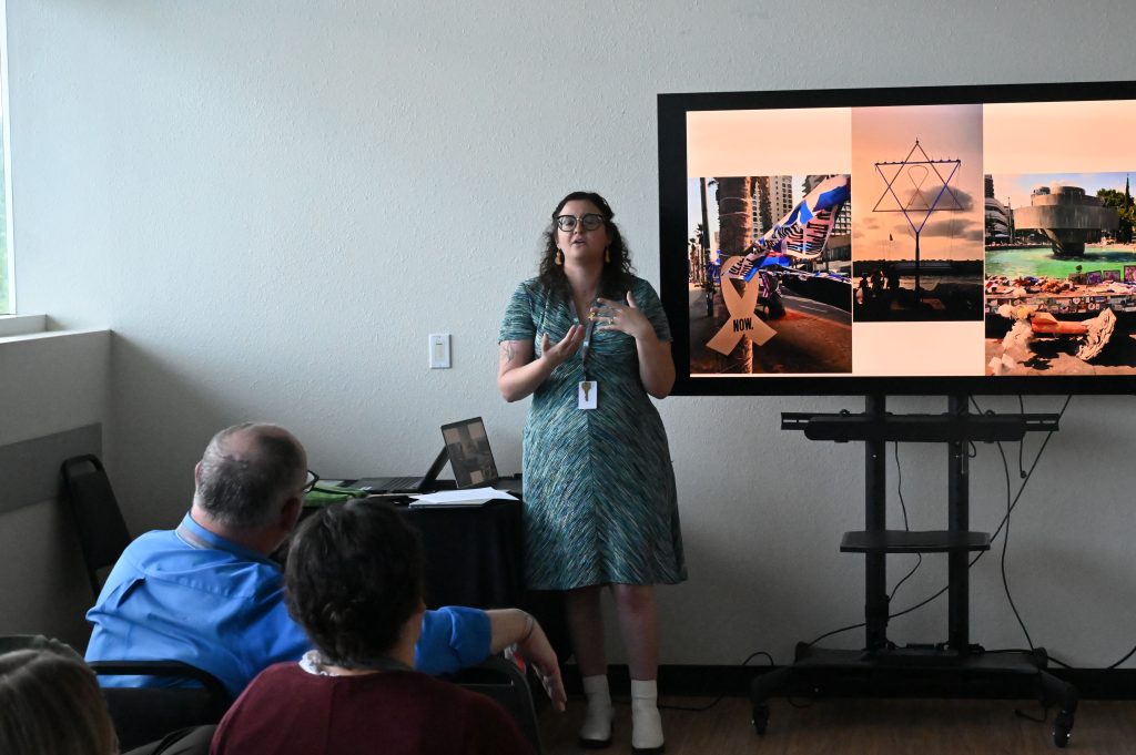 a woman stands next to a slideshow and speaks to a group of people