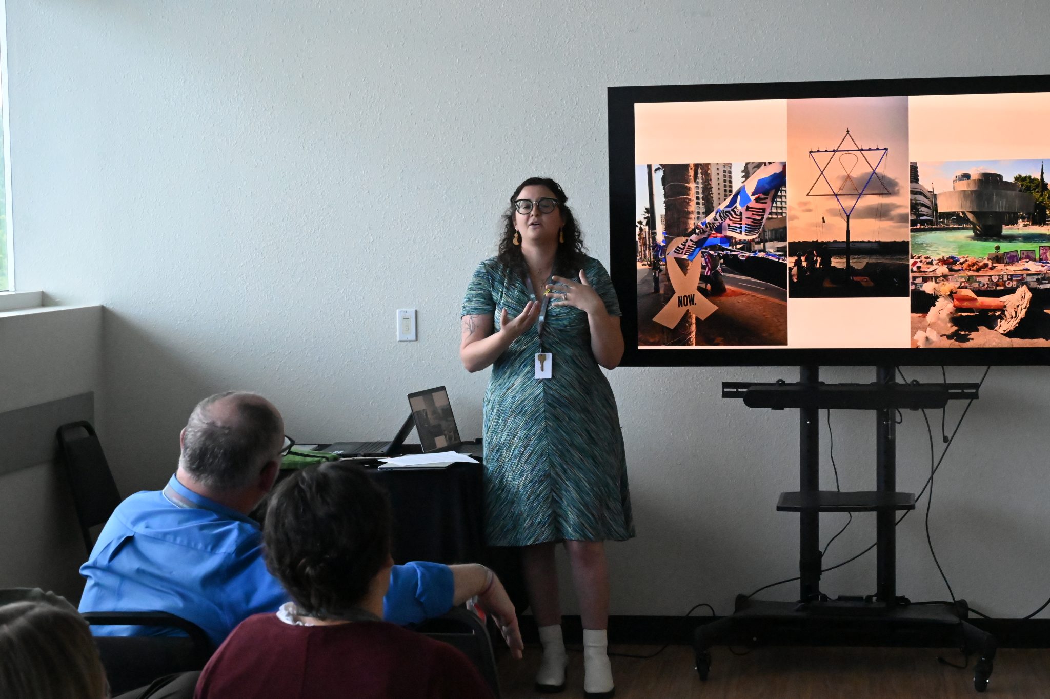 a woman stands next to a slideshow and speaks to a group of people