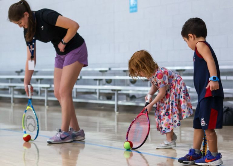two young children play with tennis racquets alongside an adult