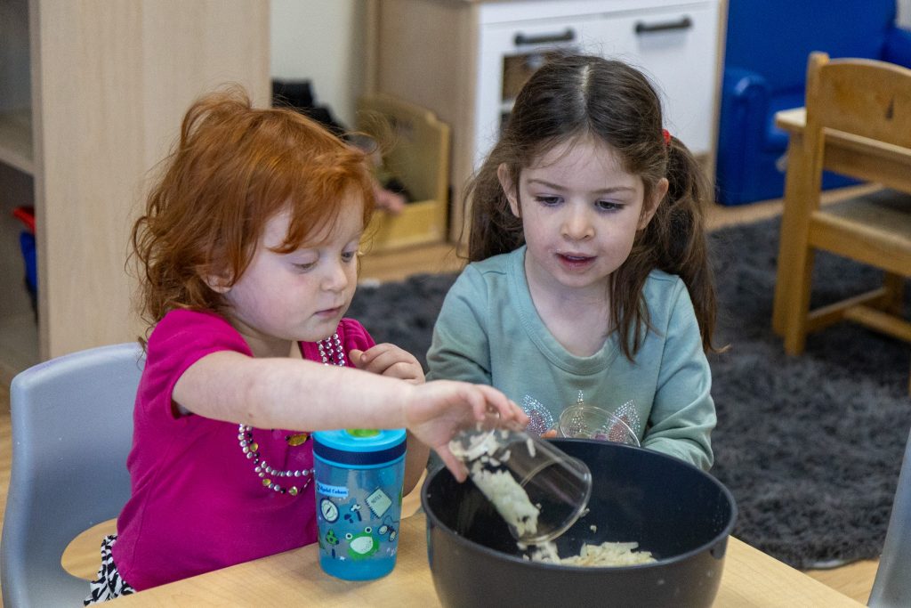 two children sit behind a bowl and pour ingrediants in